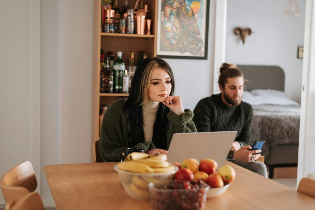 Young woman surfing internet on netbook at table with assorted fruits against boyfriend chatting on cellphone at home